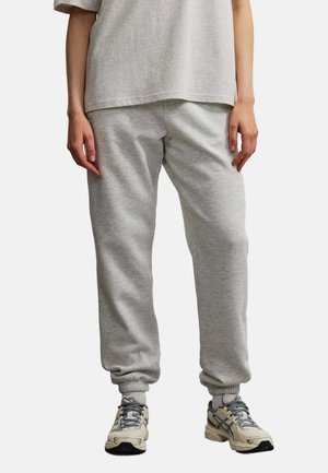 Person trägt hellgraue Sweatpants, passendes lockeres T-Shirt und beige-graue Sportschuhe, steht vor einem schlichten Hintergrund.