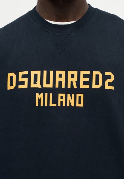 Κοντινό πλάνο σκούρου ναυτικού φούτερ με κίτρινο κείμενο "DSQUARED2 MILANO" στο στήθος σε άτομο με σκουρόχρωμο δέρμα.