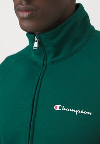 Giacca verde con zip realizzata in tessuto morbido, dotata di colletto alto e di un logo "Champion" bianco sul petto. Hardware della cerniera visibile.