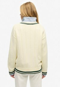 Cremfarbener gestrickter Pullover mit vertikalen Rippungen. Verfügt über einen blau-weißen gestreiften Innenkragen, grüne Akzente und einen lockeren Schnitt.