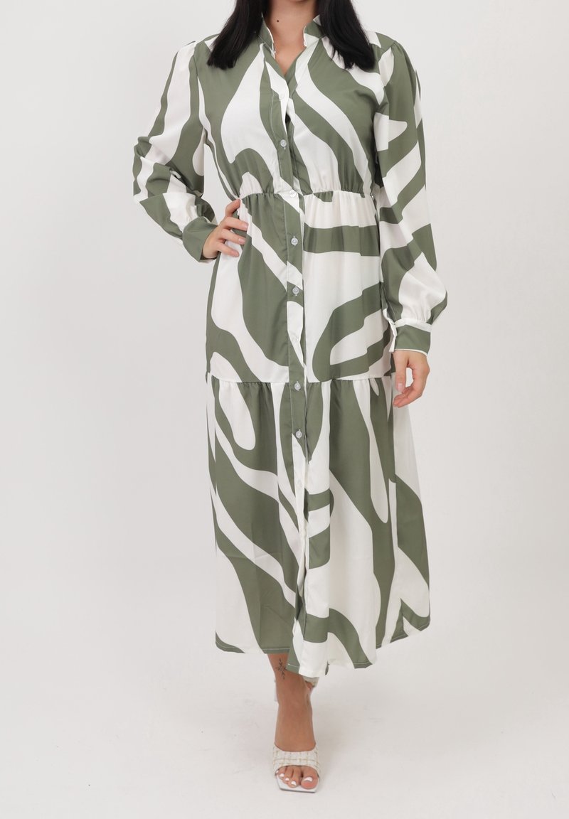 Femme portant une robe maxi à manches longues boutonnée avec de grandes rayures abstraites zébrées vertes et blanches, debout avec la main sur la hanche.