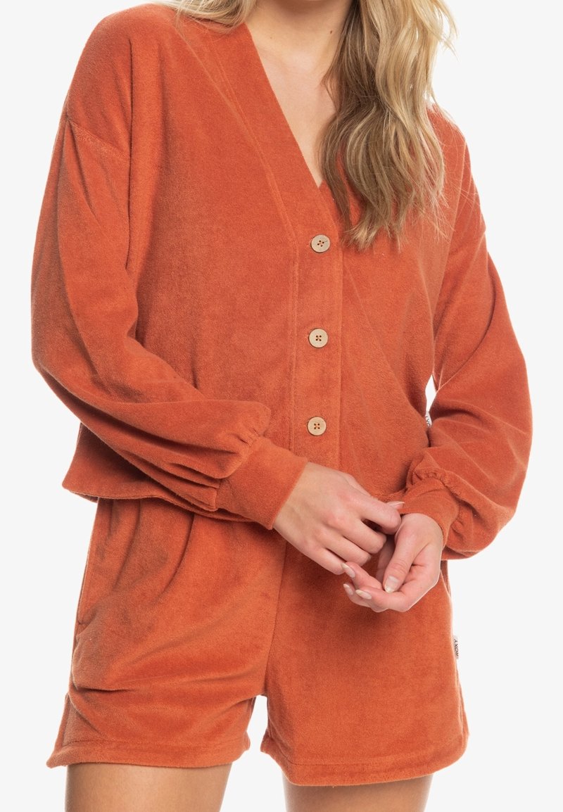Pomarańczowy komplet z polaru składający się z bluzy z dekoltem w serek zapinanej na drewniane guziki oraz rękawami z elastycznymi ściągaczami, w zestawie z dopasowanymi szortami.