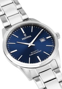 Montre en acier inoxydable argenté avec un cadran bleu marine. Elle possède des index et des aiguilles argentés, une fenêtre de date à 3 heures, et un mouvement à quartz.