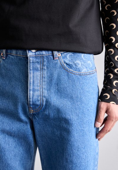 Ljusblå denimjeans med orange sömmar, med subtila mönstrade fickor. Bärs med en svart topp och dekorativa långa ärmar.