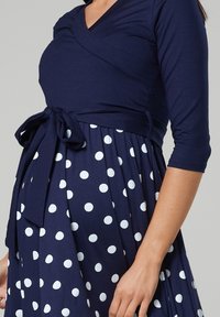 Mörkblå omlottklänning med trekvartsärmar, som har ett knytband i midjan och en kjol prydd med vita prickar. Mjuk tygtextur.
