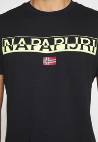Camiseta de algodón negra con un logo amarillo de "NAPAPIJRI" y un pequeño gráfico de la bandera de Noruega. Cuello redondo, mangas cortas, textura suave.