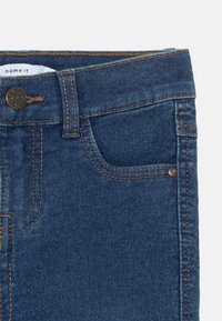 Jeans en denim bleu foncé avec une fermeture à bouton, des passants de ceinture et une poche avant avec des détails de couture contrastants. Texture lisse.