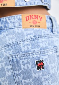Pantalones cortos de mezclilla azul claro con una textura estampada, que cuentan con una etiqueta de cuero marrón clara con la marca "DKNY" y un logo bordado "RNY" en rojo y negro.