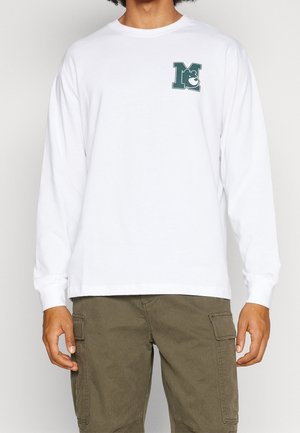 Person trägt ein weißes Langarmshirt mit einem grünen "M" und einem Bärenlogo auf der Brust, kombiniert mit olivgrünen Cargohosen.
