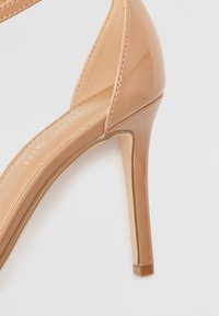 Nude spalvotos patentinės odos aukštakulniai sandalai su elegantišku dizainu, turintys smailų pirštą ir ploną stiletą, suteikiantys išskirtinį išvaizdą.