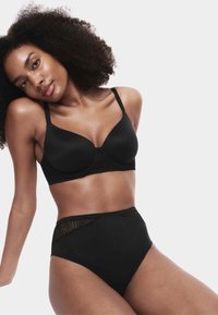 Frau mit natürlichen lockigen Haaren, die einen schwarzen Bügel-BH und hochgeschnittene Slips mit Spitzen-Details trägt und vor einem weißen Hintergrund posiert.