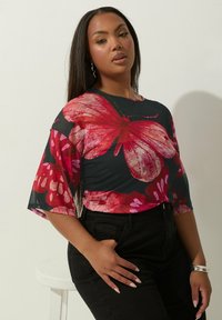 Musta korkea vyötärönhousut yhdistettynä läpikuultavaan mustaan puseroon, jossa on suuria punaisia ja vaaleanpunaisia perhoskuvioita. Puserossa on leveät hihat.