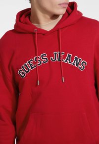 Červená mikina s kapucňou vyrobená z bavlny, s oblúkovým logom "GUESS JEANS" v čiernej a bielej výšivke. Obsahuje prednú kapsu a kapucňu so šnúrkami.