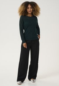 Långärmad mörkgrön spetsblus med ett strukturerat blommönster, parat med svarta vida byxor och silverfärgade skor, vilket skapar en kontrasterande look.