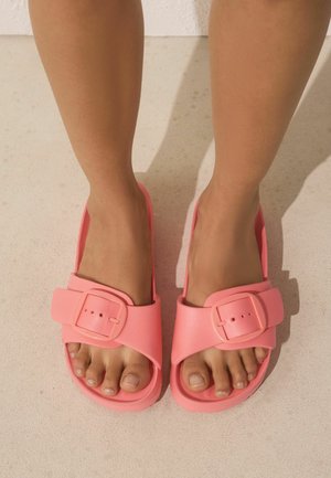 Voeten met felroze instapsandalen met grote gespbanden die op een lichtbeige oppervlak staan.