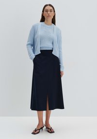 Cardigan en tricot bleu clair porté sur un pull bleu clair, associé à une jupe longue marine avec une fente à l'avant. Des sandales à talons noires complètent le look.