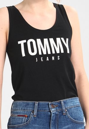 Kobieta ubrana w czarną, bezrękawnikową bluzkę z napisem "TOMMY JEANS" oraz niebieskie dżinsy z guzikiem i zamkiem błyskawicznym.