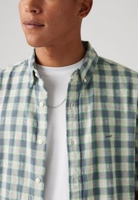 Grøn og blå ternet skjorte med knapper og krave, med hvide knapper og et Levi's logo på brystet, lagdelt over en hvid skjorte.