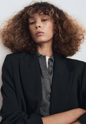 Blazer negro sobre una camiseta polo gris, con cuello y tres botones. El modelo tiene cabello rizado castaño con reflejos más claros.