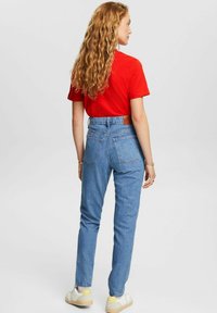 Hochgeschnittene Jeans aus Denim in Hellblau mit einem Lederpatch auf der Rückseite, kombiniert mit einem roten T-Shirt mit kurzen Ärmeln und weißen Sneakers.