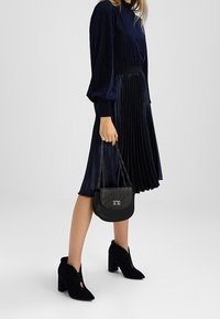 Námorně modré skládací šaty s dlouhými rukávy, úzkým pasem a černými vysokými kotníkovými botami. Doplněk: malá černá kabelka s řetízkovým popruhem.