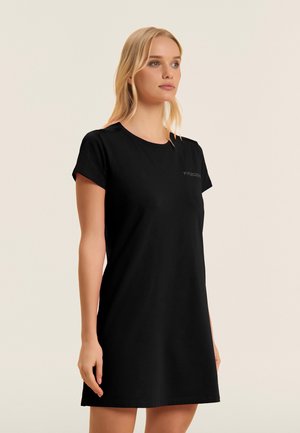 Mujer con cabello rubio, vestida con un vestido negro de manga corta, de pie frente a un fondo claro y sencillo, mirando ligeramente hacia la derecha.