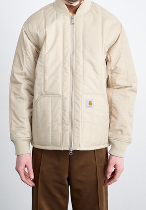Man draagt een beige gewatteerde jas met rits, geribbelde manchetten en kraag, klein Carhartt-logo op het voorvak, en een bruine broek.