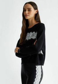 Maglione nero con un pannello in pizzo al centro, caratterizzato da una texture morbida. Abbinato a leggings neri con un accento in pizzo sul lato.