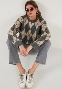 Siv in krem argyle pletena jopica z dolgimi rokavi, v kombinaciji s sivimi kavbojkami in belimi supergami. Model nosi okrogla sončna očala in uhane v obliki obroča.