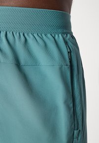 Shorts de sport teal présentant une ceinture texturée, une poche latérale zippée, et un tissu doux et lisse avec une finition légèrement réfléchissante.