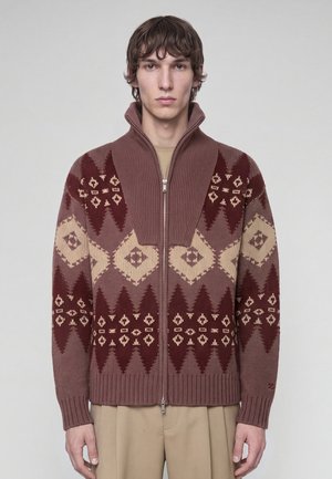 Junger Mann trägt einen braun-beigen gemusterten Strickpullover mit Reißverschluss und hohem Rippenkragen sowie beige Hosen vor einem einfarbigen Hintergrund.