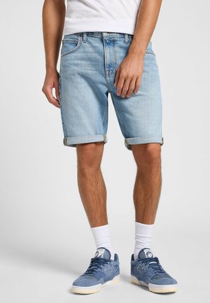 Mann trägt hellblaue hochgekrempelte Jeansshorts, weiße Socken und blaue Wildledersneaker und steht vor einem schlichten Hintergrund.