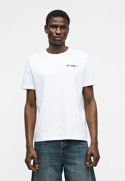 Νέος άνδρας που στέκεται, φοράει ένα λευκό T-shirt με το λογότυπο Off-White και μπλε τζιν τζιν παντελόνι, μπροστά σε απλό φωτεινό φόντο.