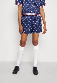 Completo sportivo in satin blu navy con motivo a fleur-de-lis bianco, con dettagli rossi e bianchi. Include pantaloni corti e calze bianche fino al ginocchio.