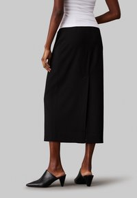 Jupe midi noire à coupe décontractée, dotée d'une fente arrière discrète et d'une texture lisse, associée à des mules noires à talon pointu.