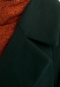 Tummanvihre ä villatakki, jossa on jämäkkä kaulus ja alla roikkuva, ruosteenvärinen teksturoitu kangas, joka tuo esiin lämpöä ja kerroksellisia suunnitteluelementtejä.