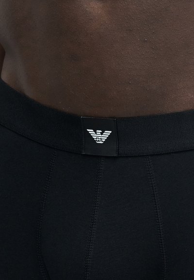 Ceinture de sous-vêtement noire avec le logo blanc Emporio Armani sur le bas du dos d'une personne à la peau foncée.