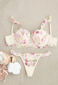 Blumen-Spitzen-BH und passender Tanga in Creme mit rosa und grünen Blumen, auf beigem Untergrund neben rosa Turnschuhen und weißen Wattepads platziert.