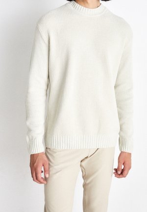 Person trägt einen weißen gerippten Strickpullover und beige Hose, steht vor einem schlichten weißen Hintergrund.
