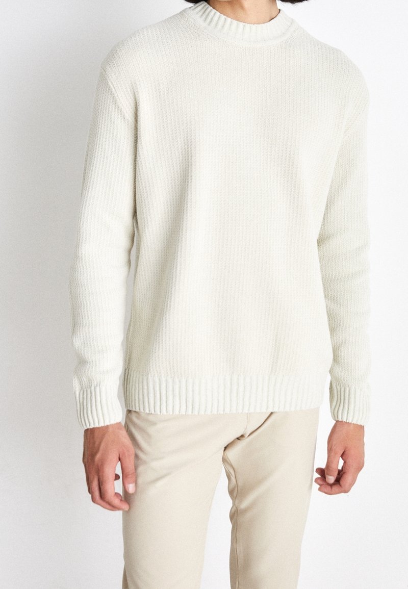 Personne portant un pull en maille côtelée blanche et un pantalon beige, debout devant un fond blanc uni.