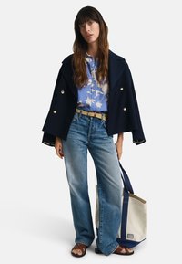 Marineblauer doppelreihiger Mantel, hellbluse Blumenshirt, hochgeschnittene hellblaue Jeans, hellbraune Sandalen und eine beigefarbene Tote-Bag mit marineblauen Akzenten.