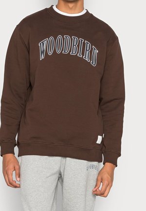Mężczyzna ubrany w brązową bluzę z napisem "WOODBIRD" oraz jasnoszare spodnie dresowe z dopasowanym małym logo "WOODBIRD".