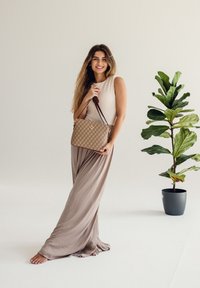 Tanova prošívaná kabelka se zlatými detaily a hnědými popruhy. Modelka má na sobě světle béžové šaty bez rukávů a stojí vedle květináče s rostlinou.