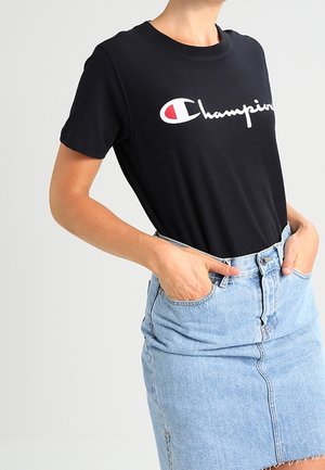 Osoba nosząca czarny t-shirt Champion wpuszczony do jasnoniebieskiej dżinsowej spódnicy z wysokim stanem, z rękami w kieszeniach spódnicy.