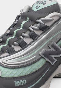 Κοντινό πλάνο σε γκρι και μέντα αθλητικό παπούτσι New Balance 1000 που δείχνει τα κορδόνια, το πλευρικό λογότυπο και τις λεπτομέρειες της υφής της σόλας.
