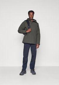 Olivengroene waterdichte jas met een capuchon, gecombineerd met donkerblauwe broek, beide met praktische zakken. Het model houdt een zwarte rugzak vast.