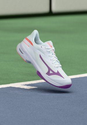 Chaussure de sport blanche avec des accents violets et orange planant au-dessus de la surface d'un court de tennis près d'une ligne blanche de délimitation.