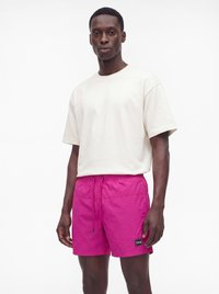 Jonge man staand, draagt een effen off-white t-shirt en felroze shorts met een trekkoord tegen een witte achtergrond.