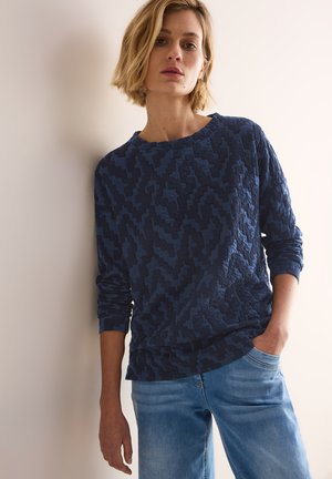 Sweatshirt texturé bleu marine avec un motif géométrique, col rond et manches longues, associé à un jean en denim bleu clair.