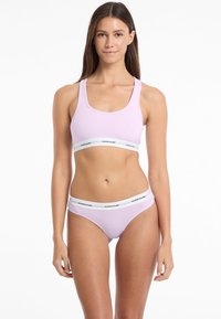 Set di reggiseno sportivo e bikini coordinato di colore lilla, con una cintura elastica bianca recante il logo "GUESS JEANS". Tessuto liscio e morbido, design semplice.